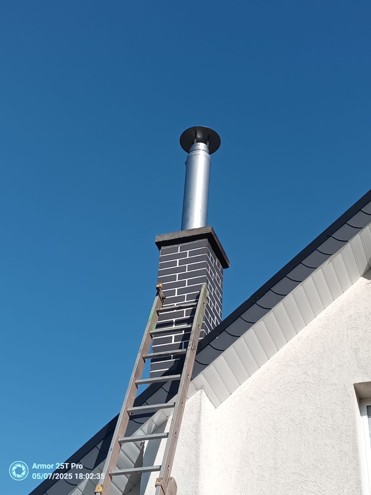 Komin z przedłużeniem ze stali nierdzewnej, drabina oparta o ceglany komin na tle błękitnego nieba. Widok z dołu na dom z białą elewacją i szarym dachem.