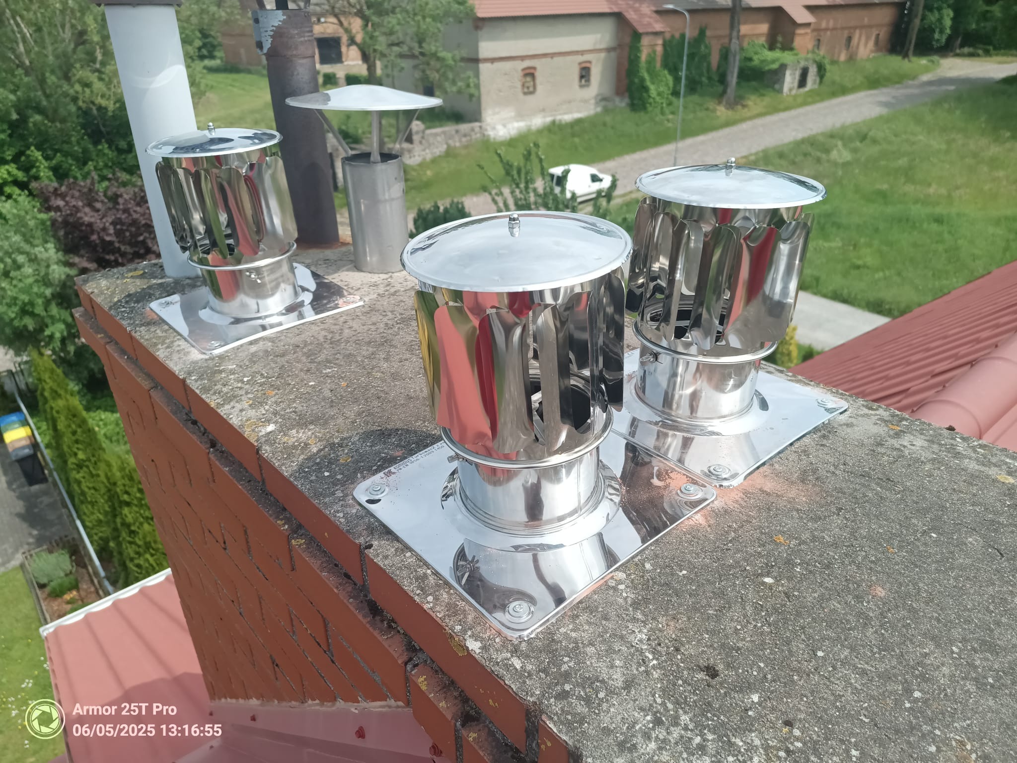 Błyszczące, metalowe nasady kominowe, typu turbowent, zamontowane na ceglanym kominie z szarą, betonową czapą, na tle zieleni i budynków. Widok z góry.