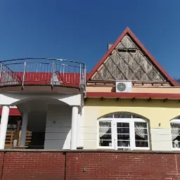 Budynek z czerwonym dachem, balkonem z metalową balustradą, oknami łukowymi i drewnianym szczytem z dwoma klimatyzatorami.