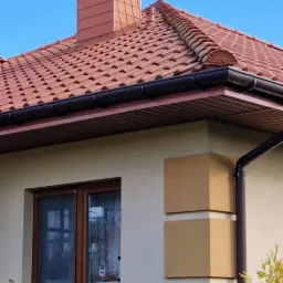 Fragment domu jednorodzinnego z czerwonym dachem ceramicznym, brązowymi rynnami i kominem z cegły klinkierowej, widok z dołu na tle błękitnego nieba.