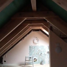 Firma Handlowo Usługowa Łukasz Jarząbek - Poddasze w trakcie remontu z widoczną konstrukcją dachu, ścianą z częściowo usuniętą starą farbą, drabiną, zwisającym przewodem elektrycznym i narzędziami na podłodze.