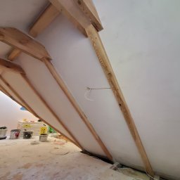 Firma Handlowo Usługowa Łukasz Jarząbek - Poddasze w trakcie prac wykończeniowych: widoczne drewniane elementy konstrukcyjne dachu, ocieplone skośne ściany, kable elektryczne wystające ze ściany, oraz narzędzia i puszki z farbą na podłodze.