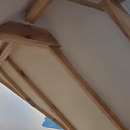 Skośny sufit poddasza z drewnianą konstrukcją nośną, częściowo pomalowany na biało, widoczne fragmenty niepomalowanej ściany.