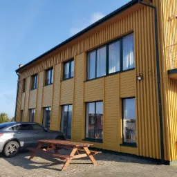 Dwukondygnacyjny budynek o żółtej elewacji z pionowymi panelami, ciemnozielonymi ramami okiennymi, szary samochód i drewniany stół piknikowy na brukowanym podjeździe w słoneczny dzień.