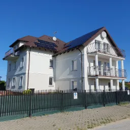 Trzykondygnacyjny dom z białymi ścianami, brązowym dachem i panelami słonecznymi, widziany zza czarnego ogrodzenia.