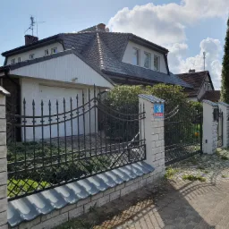 Dom jednorodzinny z ciemną dachówką, jasną elewacją i kutym ogrodzeniem, widziany z poziomu chodnika z kostki brukowej w słoneczny dzień.