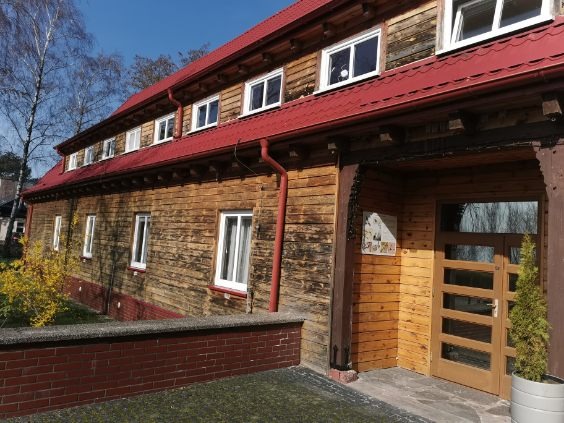 Drewniany budynek z czerwonym dachem, widoczne okna, rynny i wejście z przeszklonymi drzwiami, drzewa w tle.