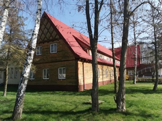 Drewniany budynek z czerwonym dachem, widoczny przez pnie drzew, z fragmentem drugiego budynku w tle.