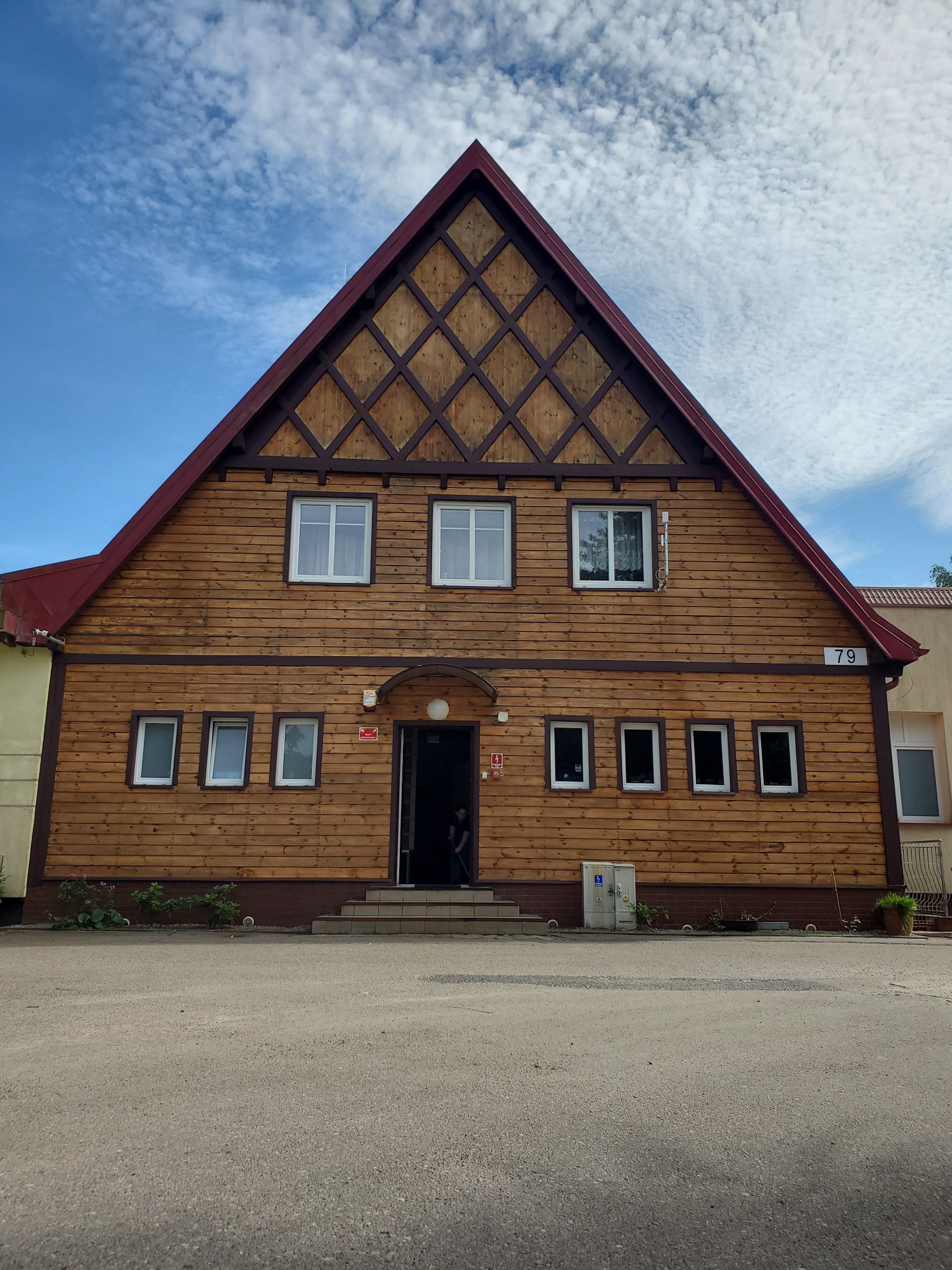 Drewniana elewacja budynku z numerem 79, z widocznym wzorem kratownicy pod dachem i kilkoma oknami, na tle błękitnego nieba z obłokami.