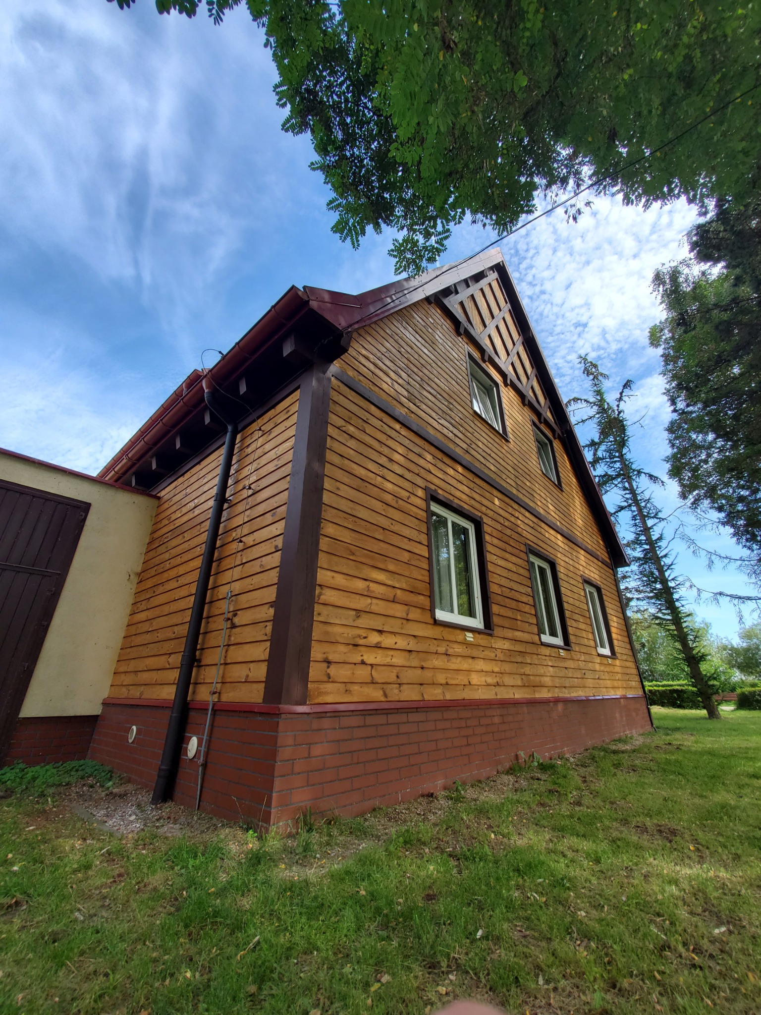 Drewniana elewacja domu z brązowymi elementami wykończeniowymi, widok z dołu na tle błękitnego nieba i zieleni drzew.