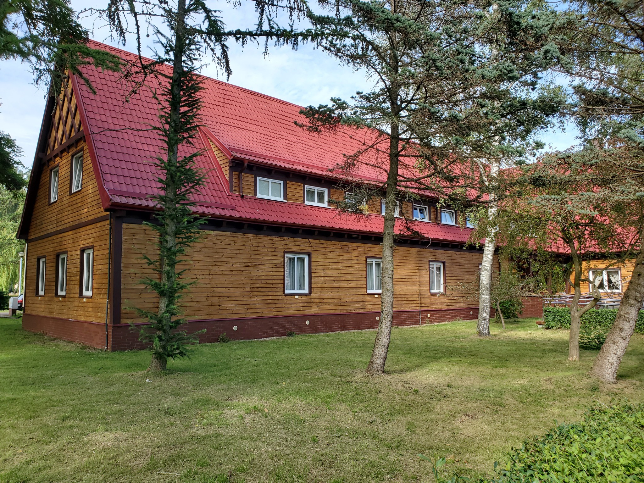 Drewniany budynek z czerwonym dachem widziany z perspektywy trawnika, częściowo zasłonięty przez drzewa iglaste i brzozy.