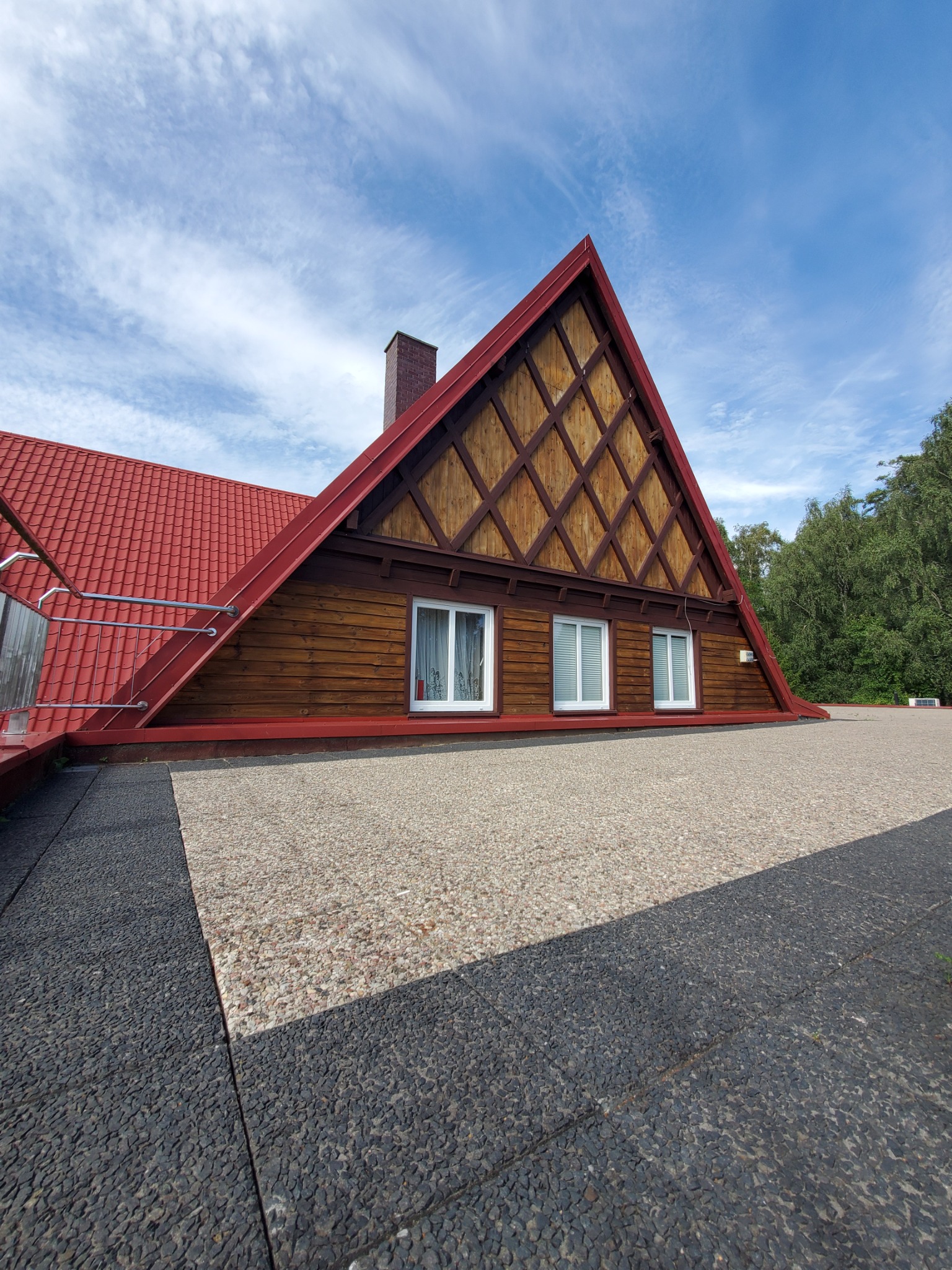 Dom z trójkątną elewacją szczytową, drewnianą boazerią w romby, czerwonym dachem i kominem, widok z góry na płaski dach pokryty żwirem i kostką brukową.