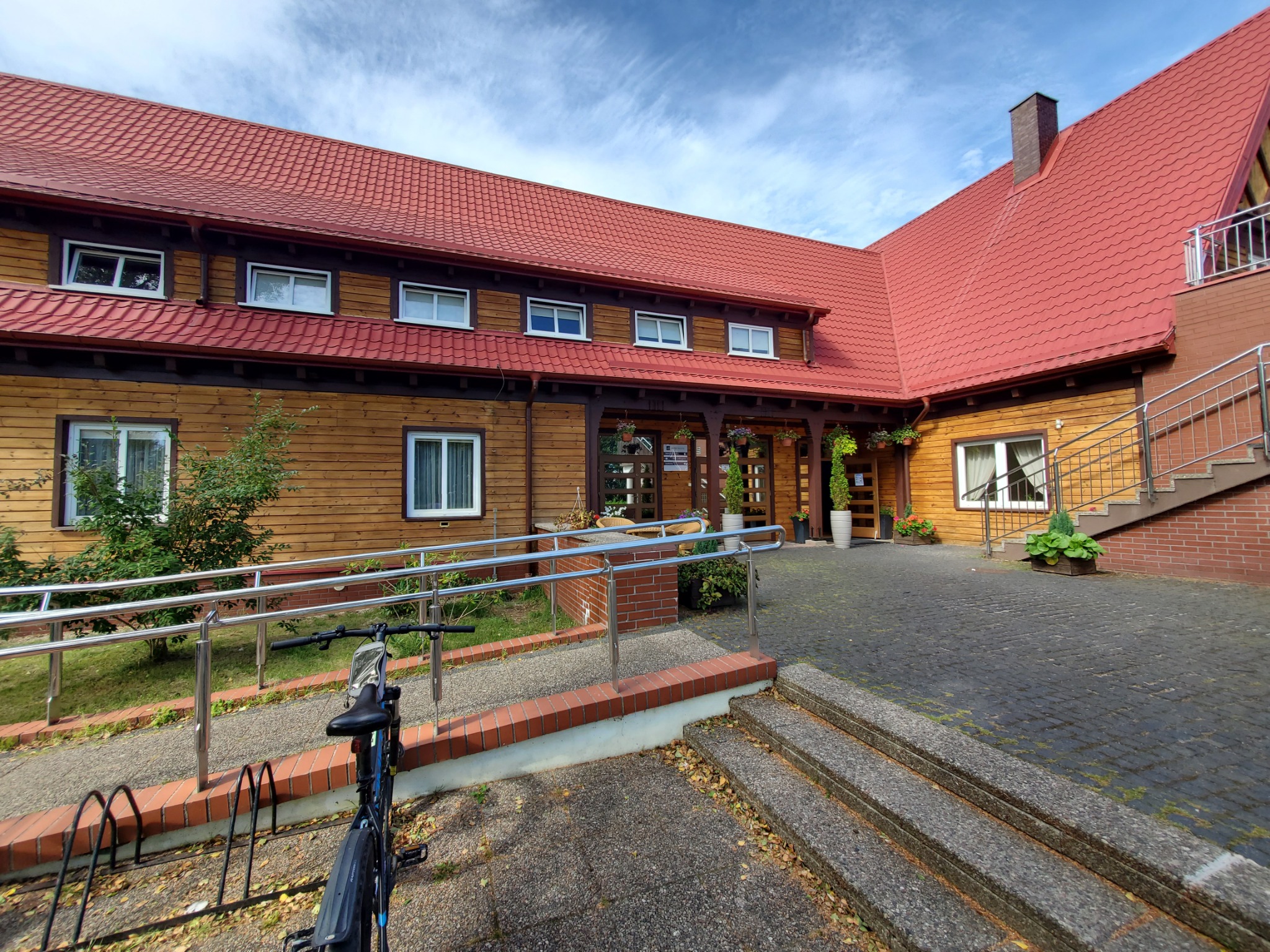 Drewniany budynek z czerwonym dachem, widok z zewnątrz, rower zaparkowany przed budynkiem, kostka brukowa na podjeździe.