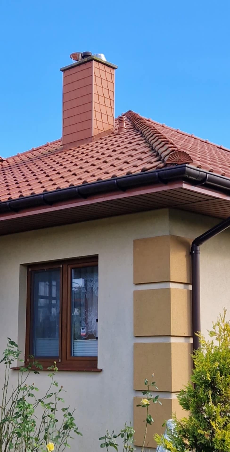 Fragment domu jednorodzinnego z czerwonym dachem ceramicznym, brązowymi rynnami i kominem z cegły klinkierowej, widok z dołu na tle błękitnego nieba.