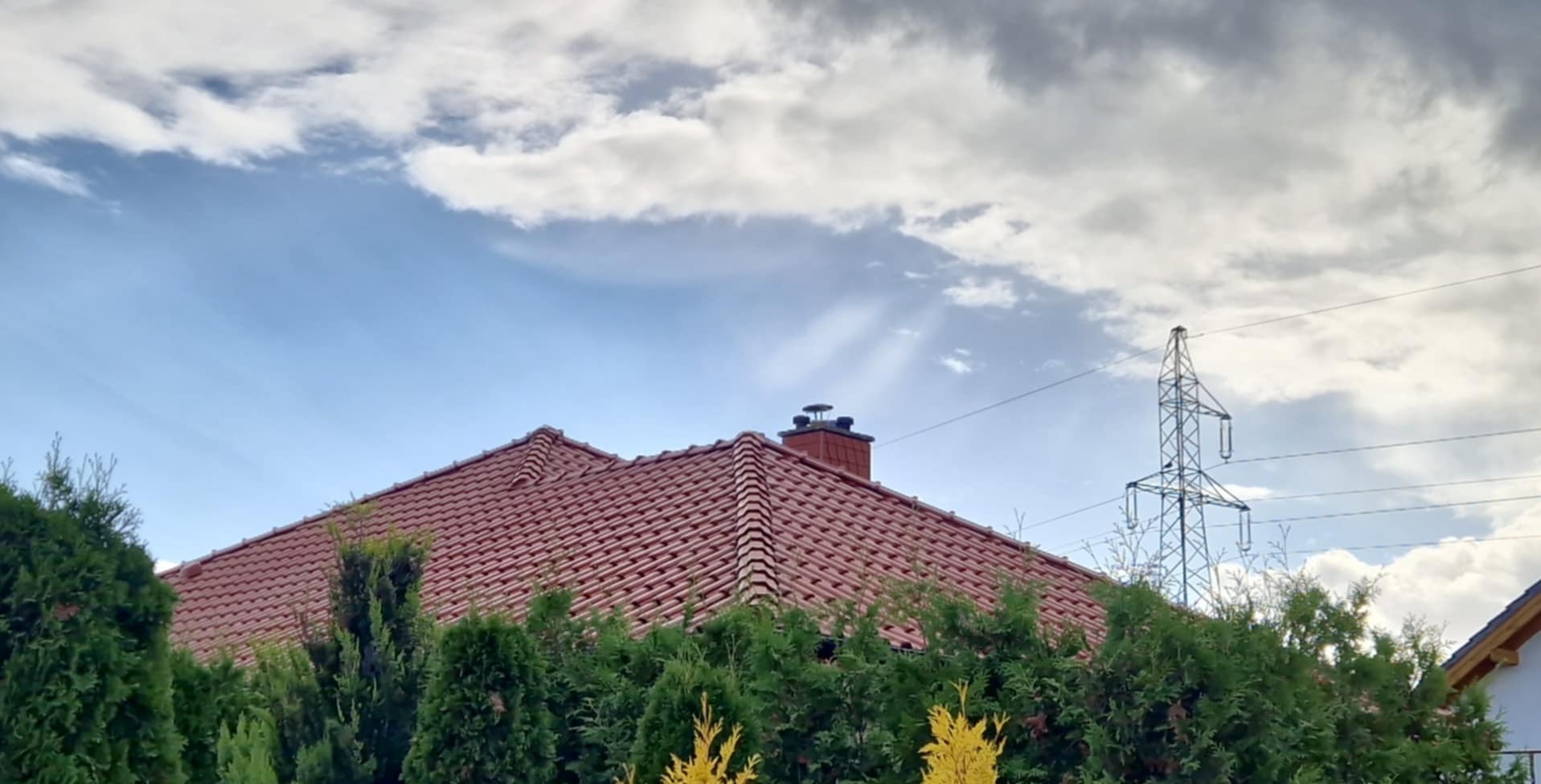 Czerwony dach z kominem, częściowo zasłonięty zielonymi krzewami, na tle błękitnego nieba z chmurami i słupem wysokiego napięcia.