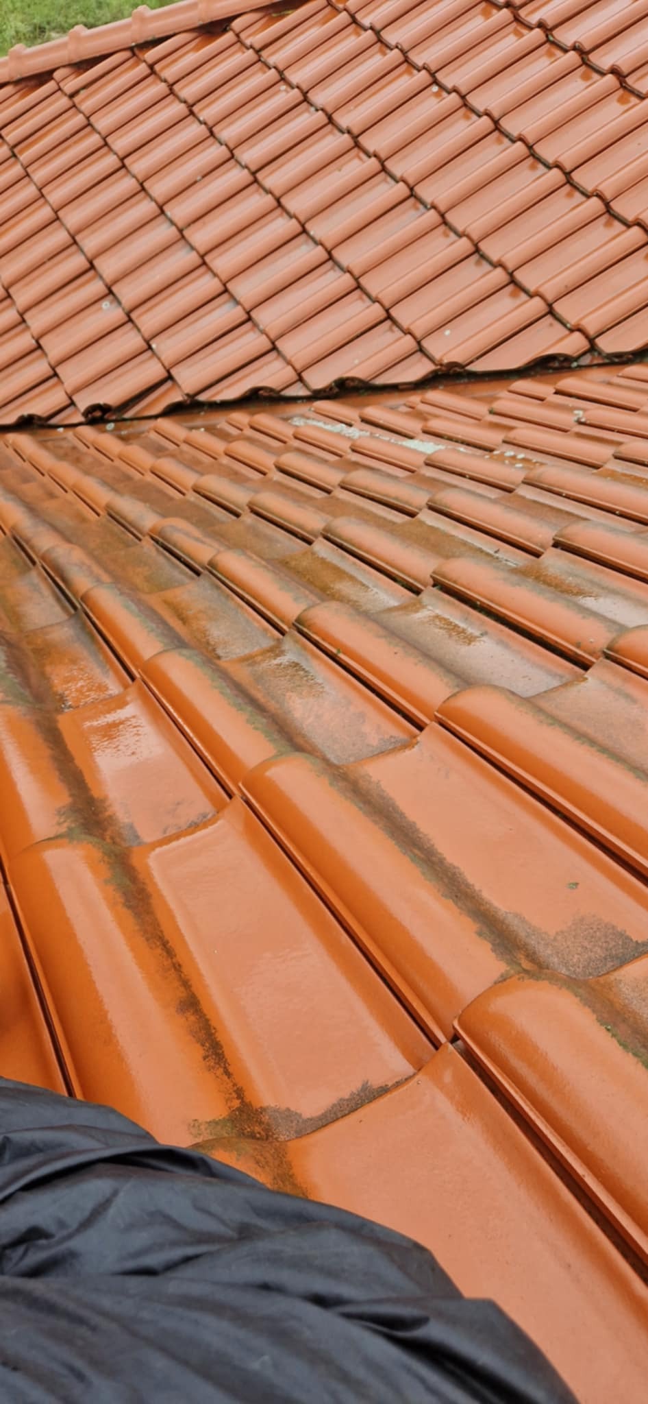 Częściowo umyty dach z pomarańczowej dachówki ceramicznej, widoczne ślady brudu i mchu, zabezpieczony folią w dolnym rogu.
