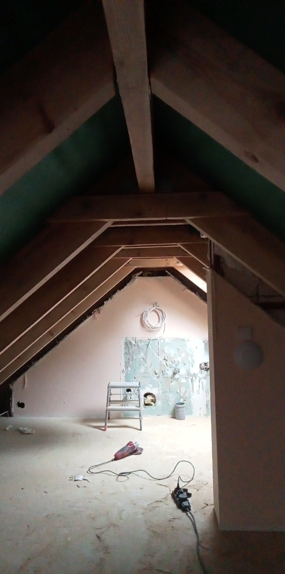 Poddasze w trakcie remontu z widoczną konstrukcją dachu, ścianą z częściowo usuniętą starą farbą, drabiną, zwisającym przewodem elektrycznym i narzędziami na podłodze.