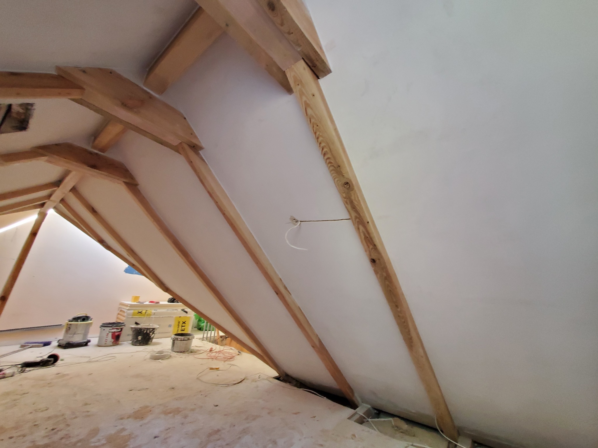 Poddasze w trakcie prac wykończeniowych: widoczne drewniane elementy konstrukcyjne dachu, ocieplone skośne ściany, kable elektryczne wystające ze ściany, oraz narzędzia i puszki z farbą na podłodze.