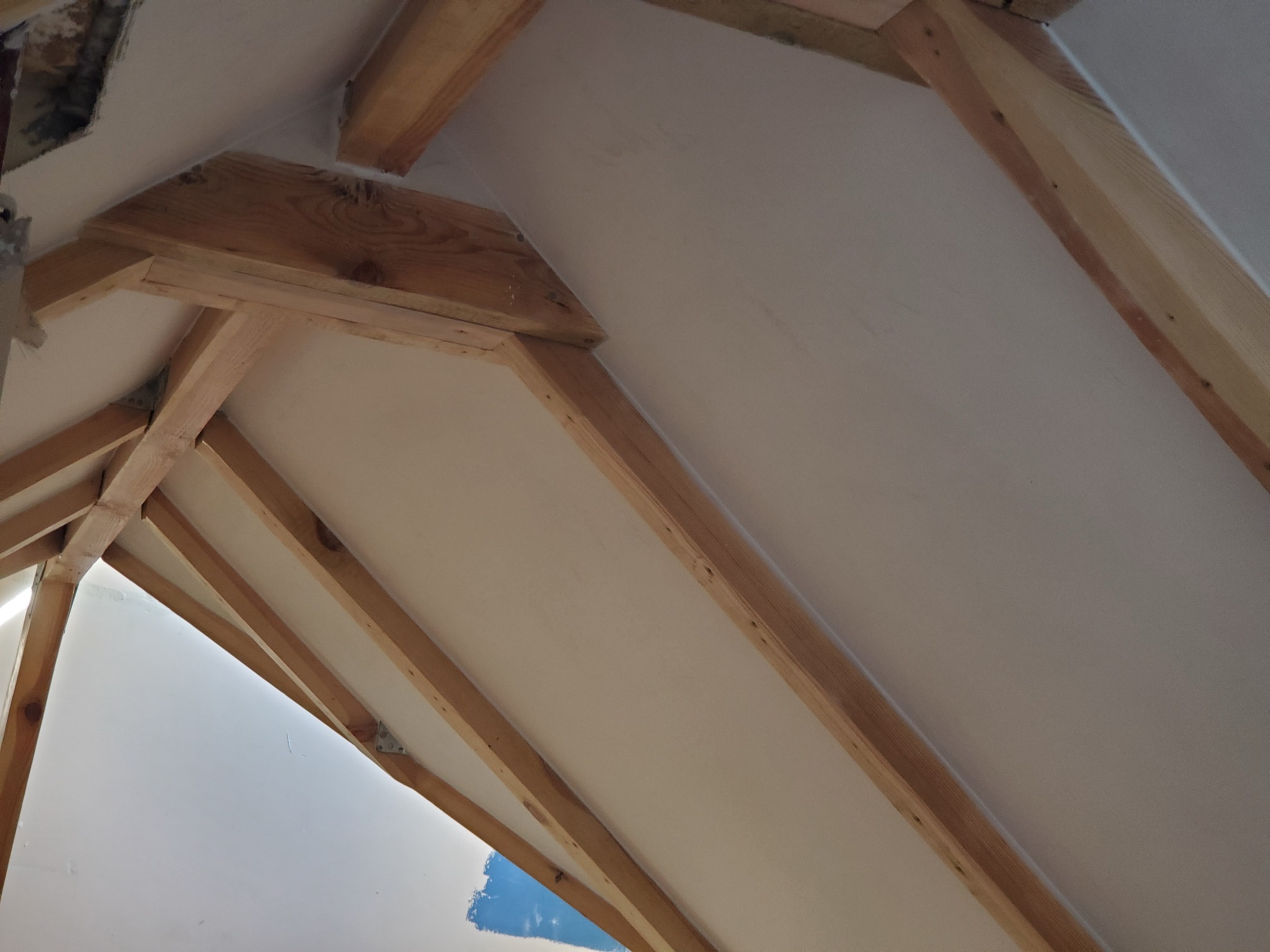 Skośny sufit poddasza z drewnianą konstrukcją nośną, częściowo pomalowany na biało, widoczne fragmenty niepomalowanej ściany.