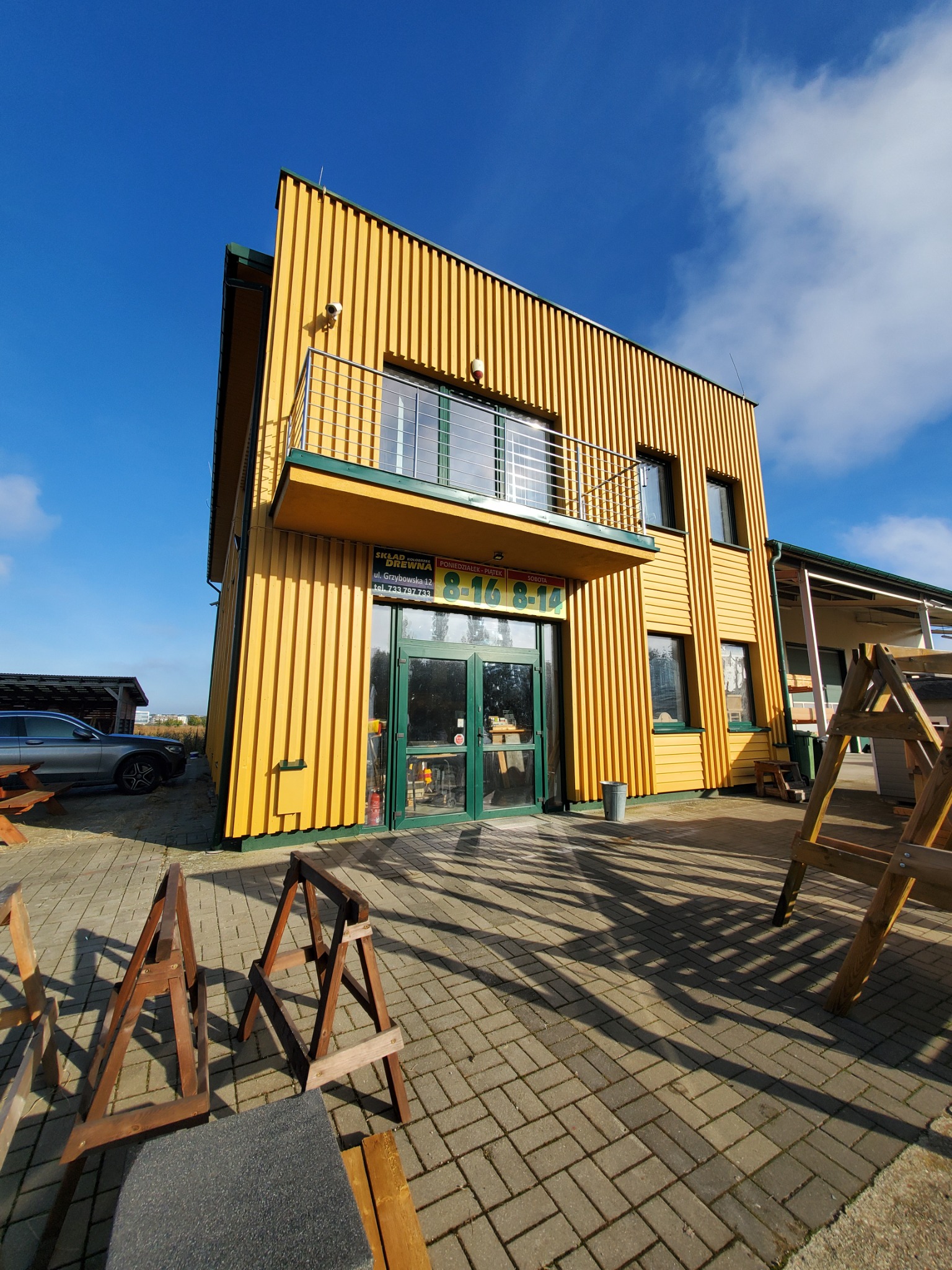 Dwukondygnacyjny budynek z elewacją z pionowych desek pomalowanych na żółto, z balkonem z metalową balustradą, zielonymi obramowaniami okien i drzwi, oraz szyldem nad wejściem, widziany z dołu,...