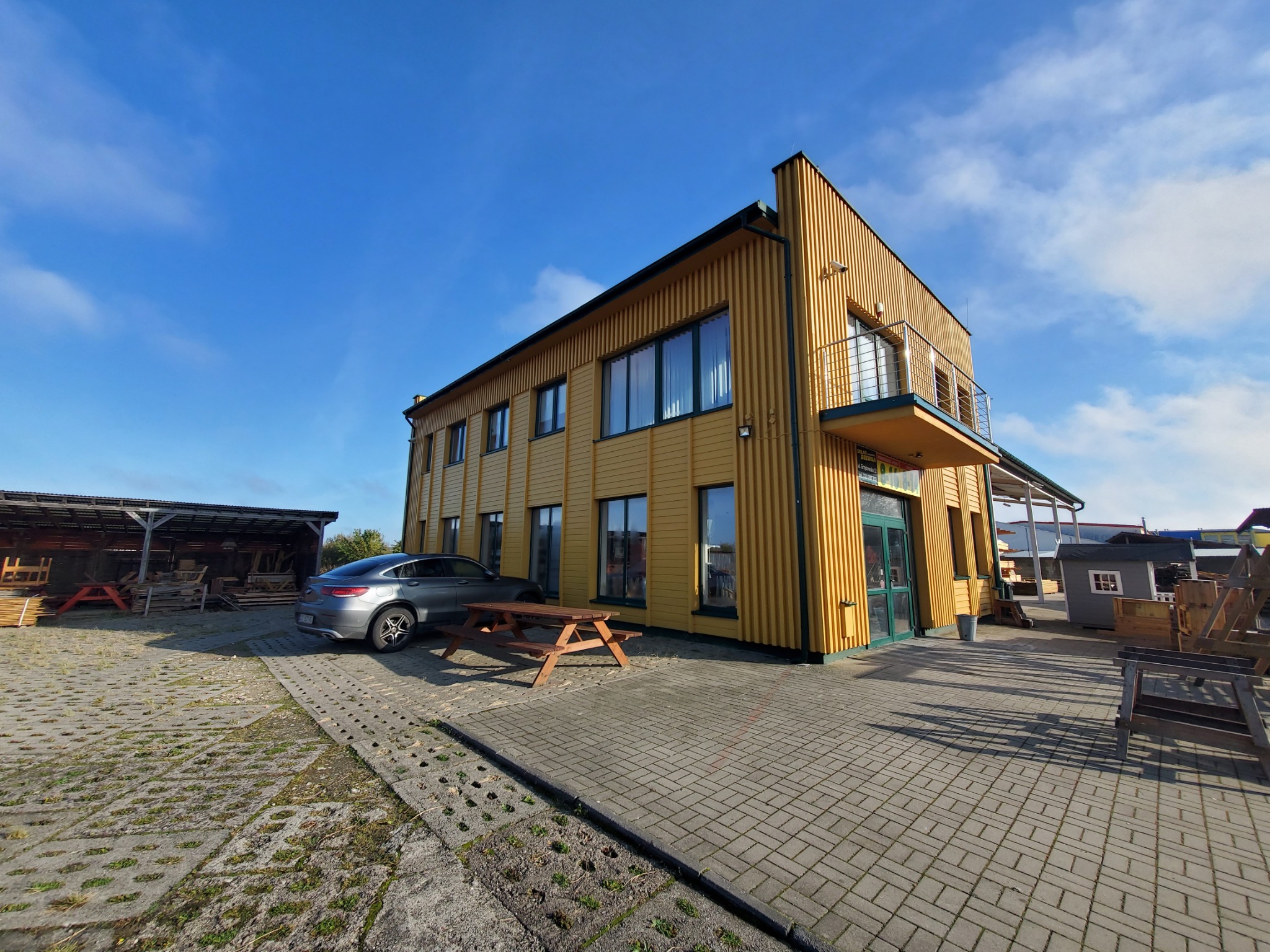 Dwukondygnacyjny budynek z żółtą elewacją z pionowych desek, balkonem i ciemnozielonymi oknami, widoczny na tle błękitnego nieba, z zaparkowanym szarym samochodem i drewnianym stołem piknikowym...