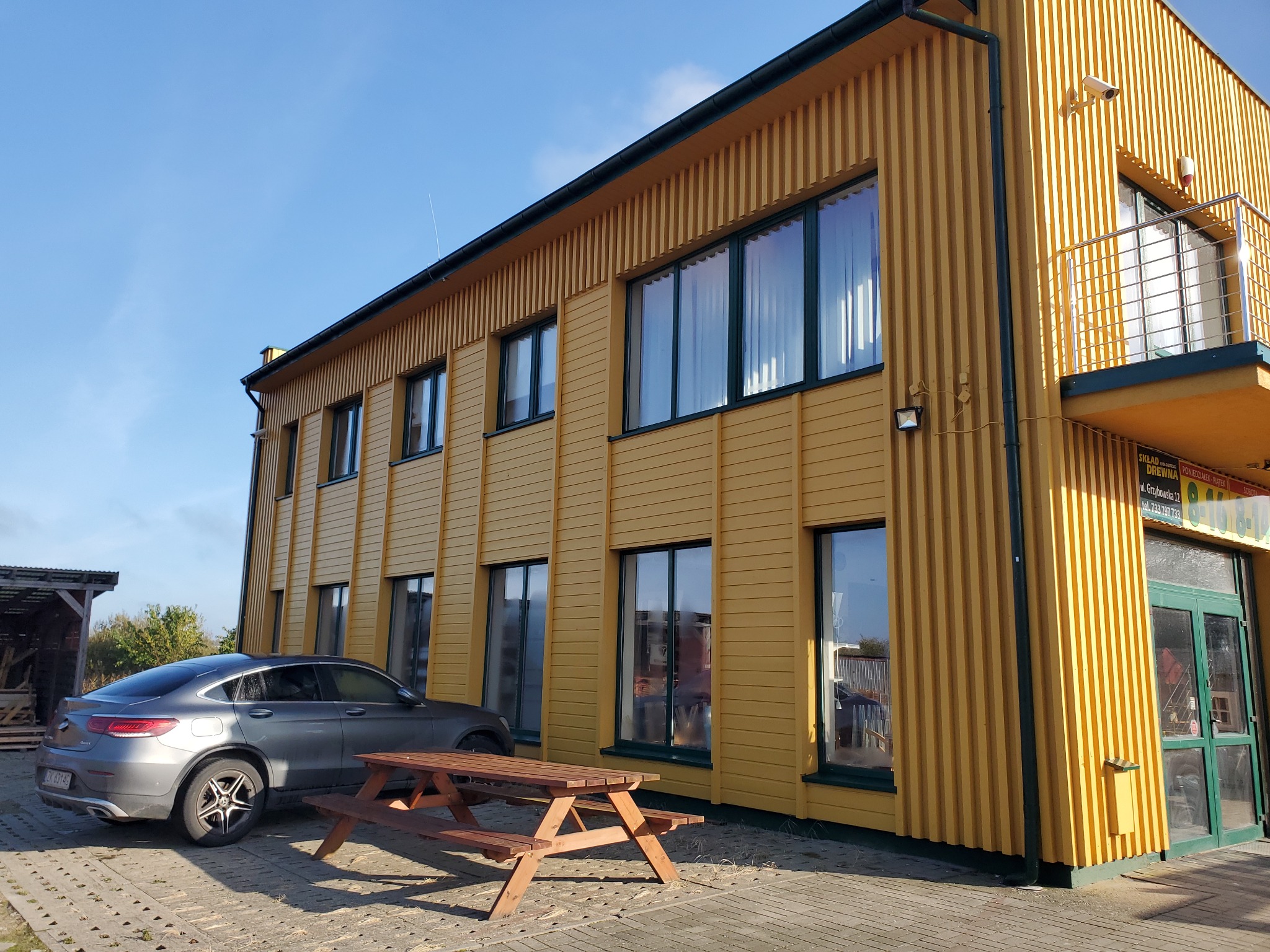 Dwukondygnacyjny budynek o żółtej elewacji z pionowymi panelami, ciemnozielonymi ramami okiennymi, szary samochód i drewniany stół piknikowy na brukowanym podjeździe w słoneczny dzień.