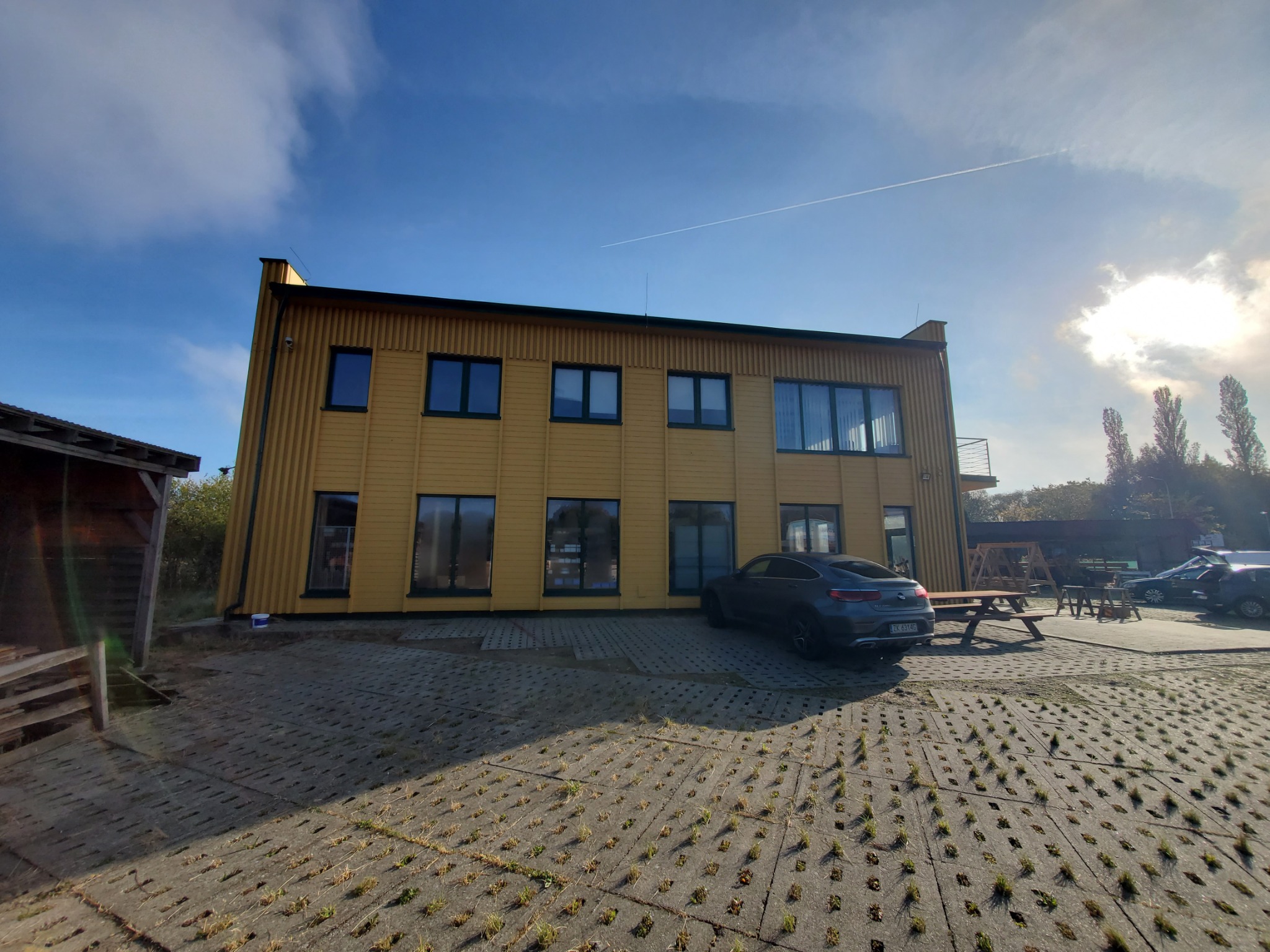Dwukondygnacyjny budynek z drewnianą elewacją w odcieniach żółci, z ciemnymi ramami okiennymi, stojący na placu wyłożonym ażurową kostką brukową, obok zaparkowany szary samochód osobowy.