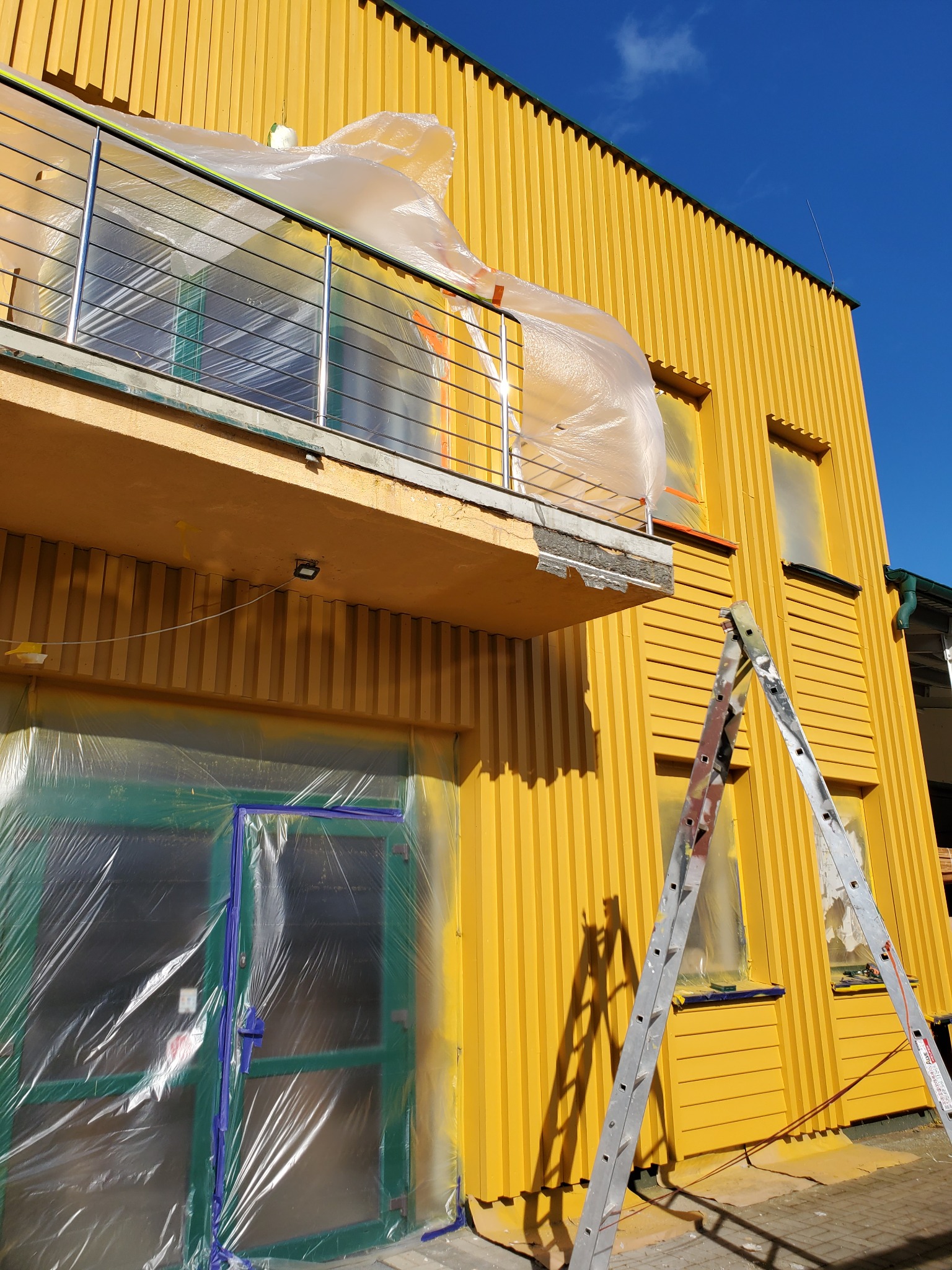 Żółta elewacja budynku w trakcie zabezpieczania folią i malowania, widoczna drabina i częściowo zakryte okna oraz balkon.