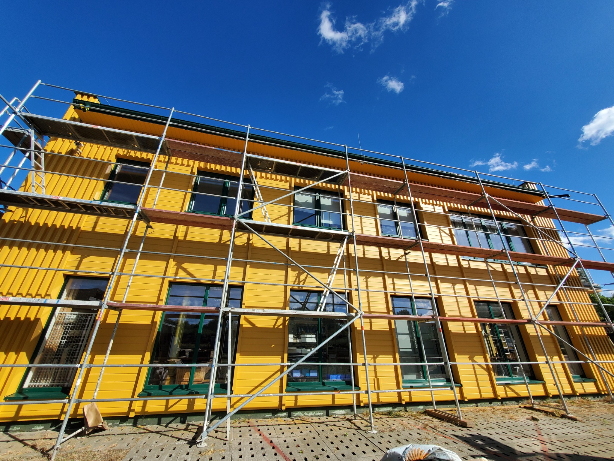 Żółta elewacja domu w trakcie prac, widoczne rusztowania, zielone ramy okienne, błękitne niebo z chmurami w tle.