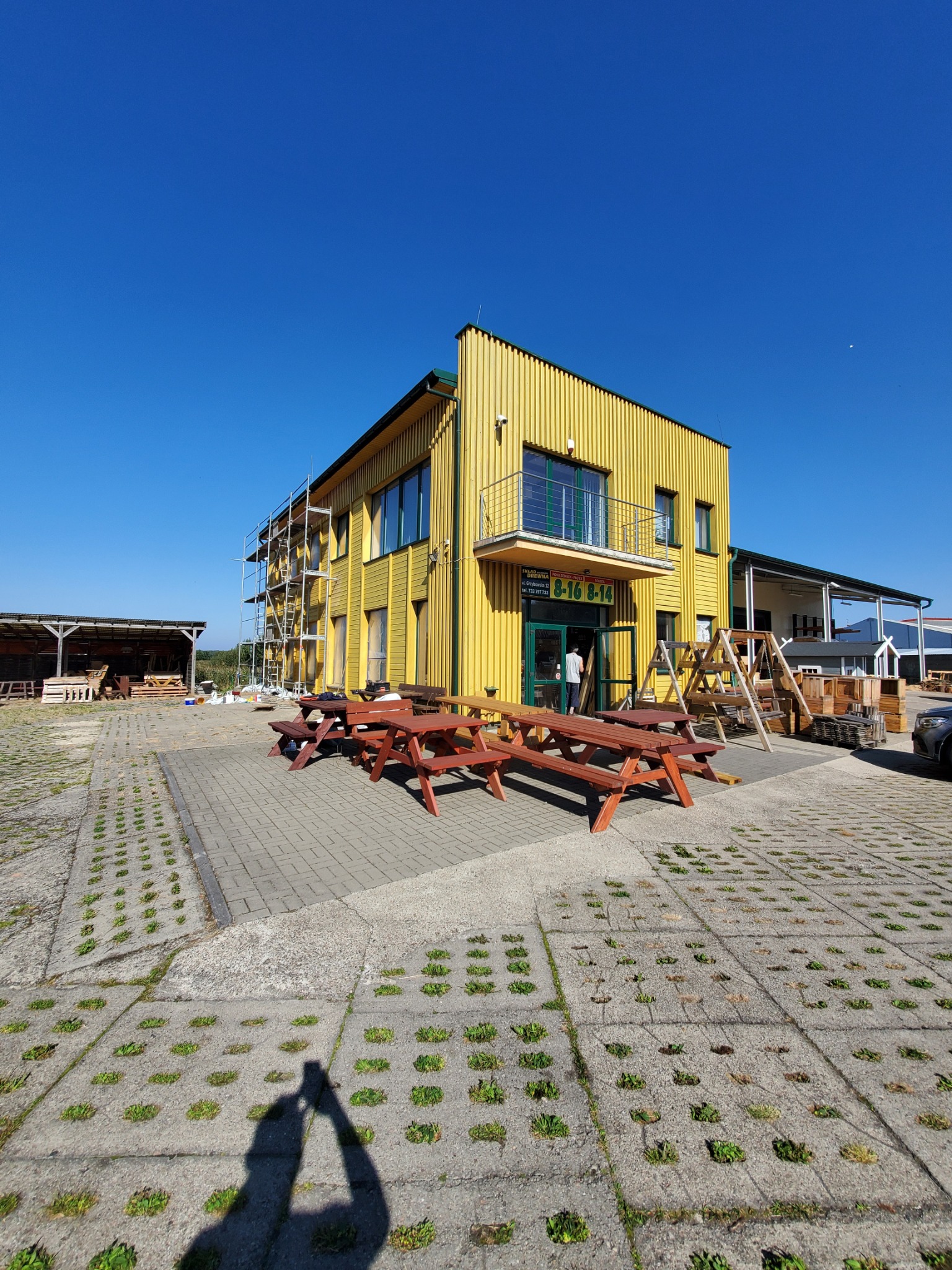 Żółty budynek z drewnianą elewacją w trakcie malowania, widoczne rusztowania, kostka brukowa z zielonymi elementami, oraz drewniane stoły piknikowe na pierwszym planie.