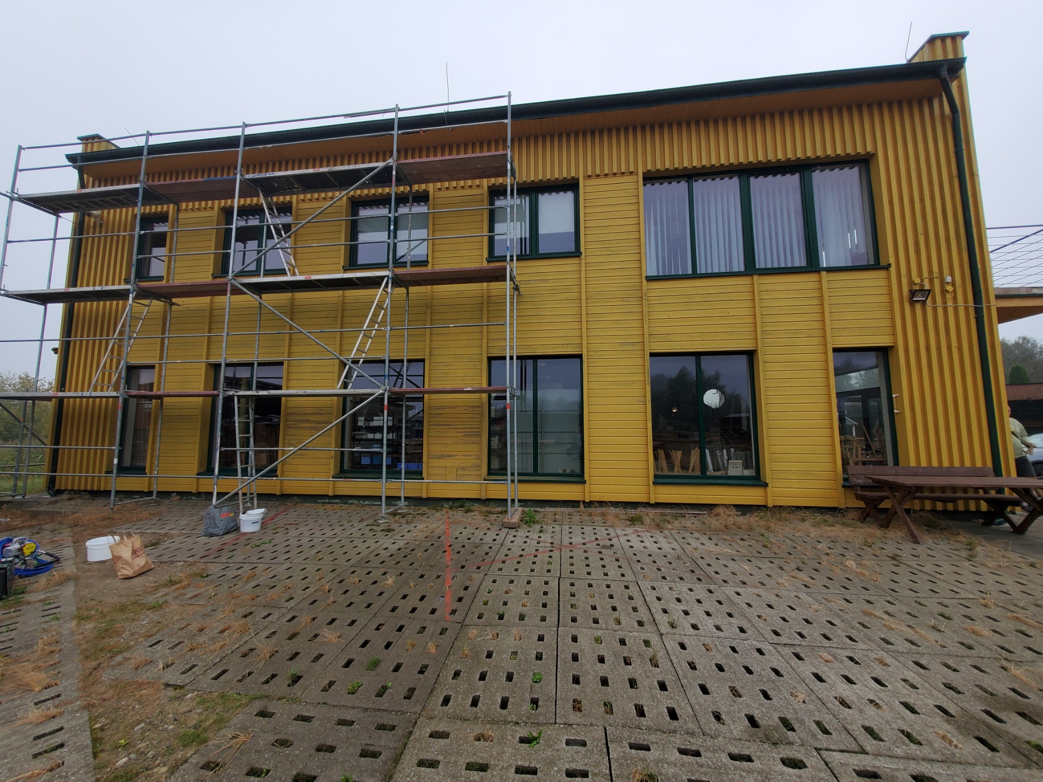 Żółta elewacja budynku z ciemnozielonymi obramowaniami okien w trakcie prac, widoczne rusztowania, wiadro z farbą i akcesoria malarskie na betonowej kostce brukowej z porostami.