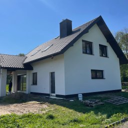 DAMIAN MĄCZKA - Nowoczesny dom jednorodzinny z białą elewacją, ciemną dachówką i czarnymi oknami, widoczny z zewnątrz w słoneczny dzień, z przygotowanym terenem wokół budynku.