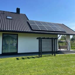 DAMIAN MĄCZKA - Biały dom jednorodzinny z ciemną dachówką i panelami słonecznymi, widok z ogrodu w słoneczny dzień.