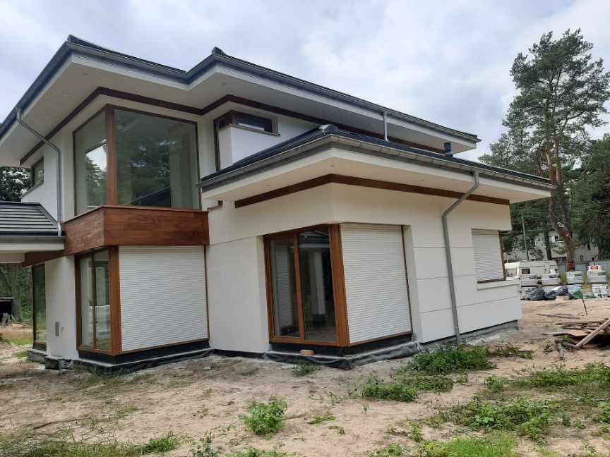 Nowoczesny dom jednorodzinny z białymi ścianami i drewnianymi elementami wykończeniowymi, częściowo zasłonięte rolety zewnętrzne, widoczne prace wykończeniowe wokół budynku.