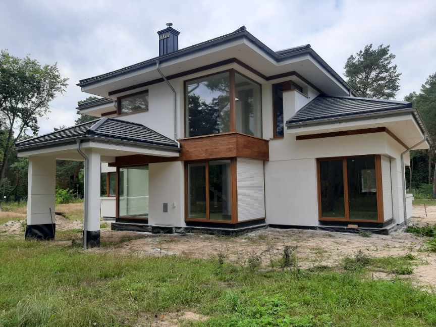 Nowoczesny dom jednorodzinny z białą elewacją, dużymi drewnianymi oknami i ciemną dachówką, otoczony zielenią.