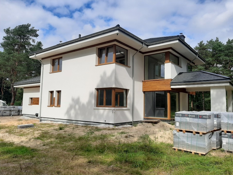 Dwukondygnacyjny dom z białym tynkiem i drewnianymi ramami okiennymi, z ciemną dachówką, otoczony zielenią, z paletami kostki brukowej w pobliżu