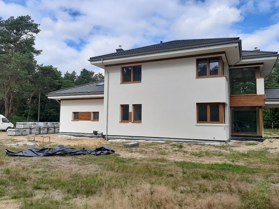 Nowoczesny dom z białą elewacją i drewnianymi oknami, widoczny w trakcie prac wykończeniowych, z materiałami budowlanymi i plandeką na trawniku.