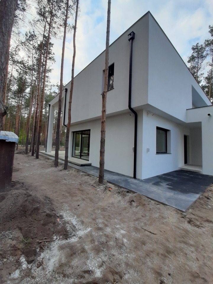 Nowoczesny, biały dom jednorodzinny z płaskim dachem, czarnymi rynnami i dużymi oknami, otoczony sosnowym lasem, z widocznym gruntem przygotowanym pod trawnik.