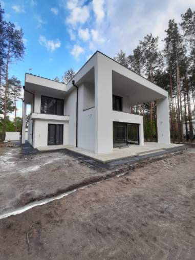 Nowoczesny, dwukondygnacyjny dom z białymi ścianami i płaskim dachem, otoczony drzewami i niezagospodarowanym terenem. Widoczne czarne rynny i okna. Dom w stylu minimalistycznym.