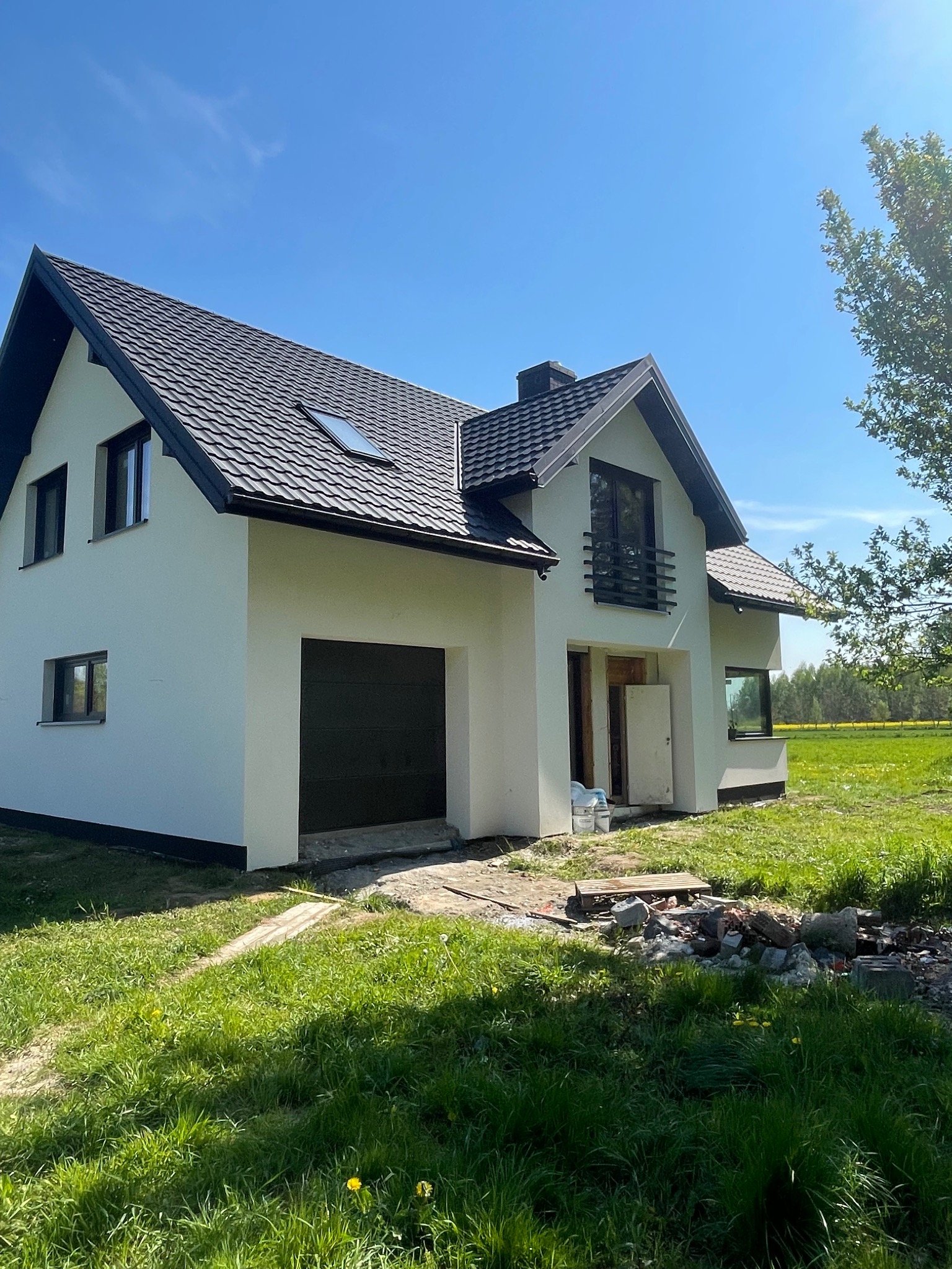 Dwupiętrowy dom jednorodzinny z ciemną dachówką i białymi ścianami, widoczne ślady prac budowlanych na trawniku, otwarte drzwi wejściowe, błękitne niebo w tle.