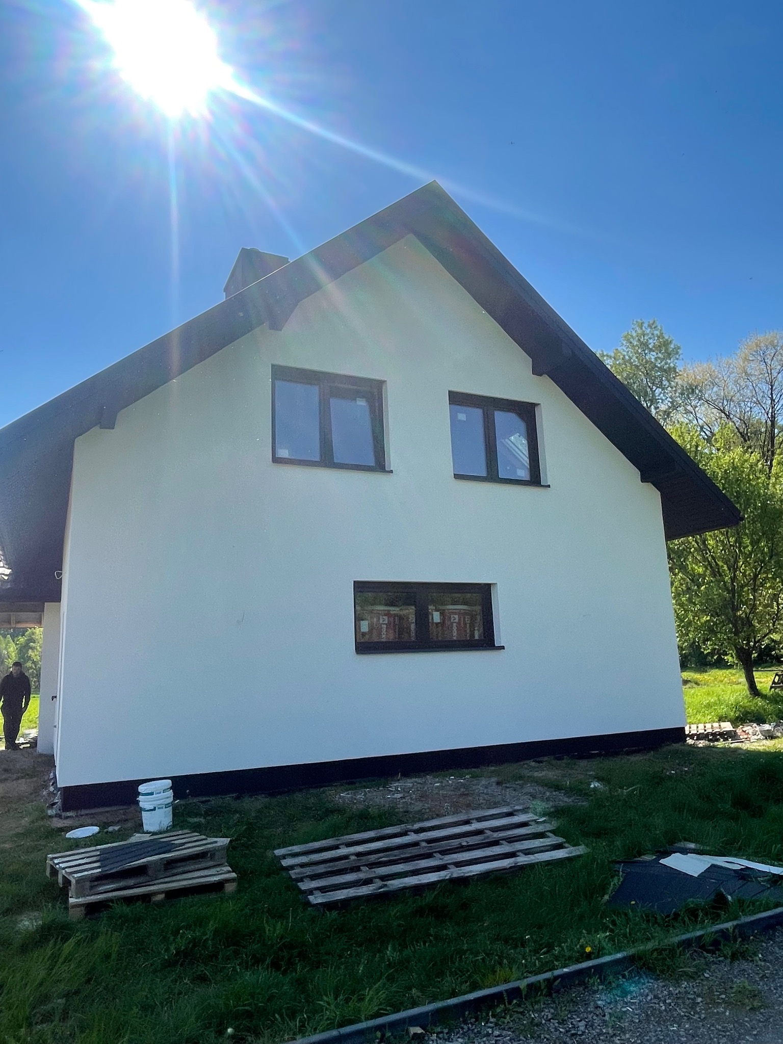 Świeżo otynkowana, biała elewacja domu jednorodzinnego z czarnymi ramami okiennymi, palety i wiadro z farbą na trawniku, widok z dołu w słoneczny dzień.