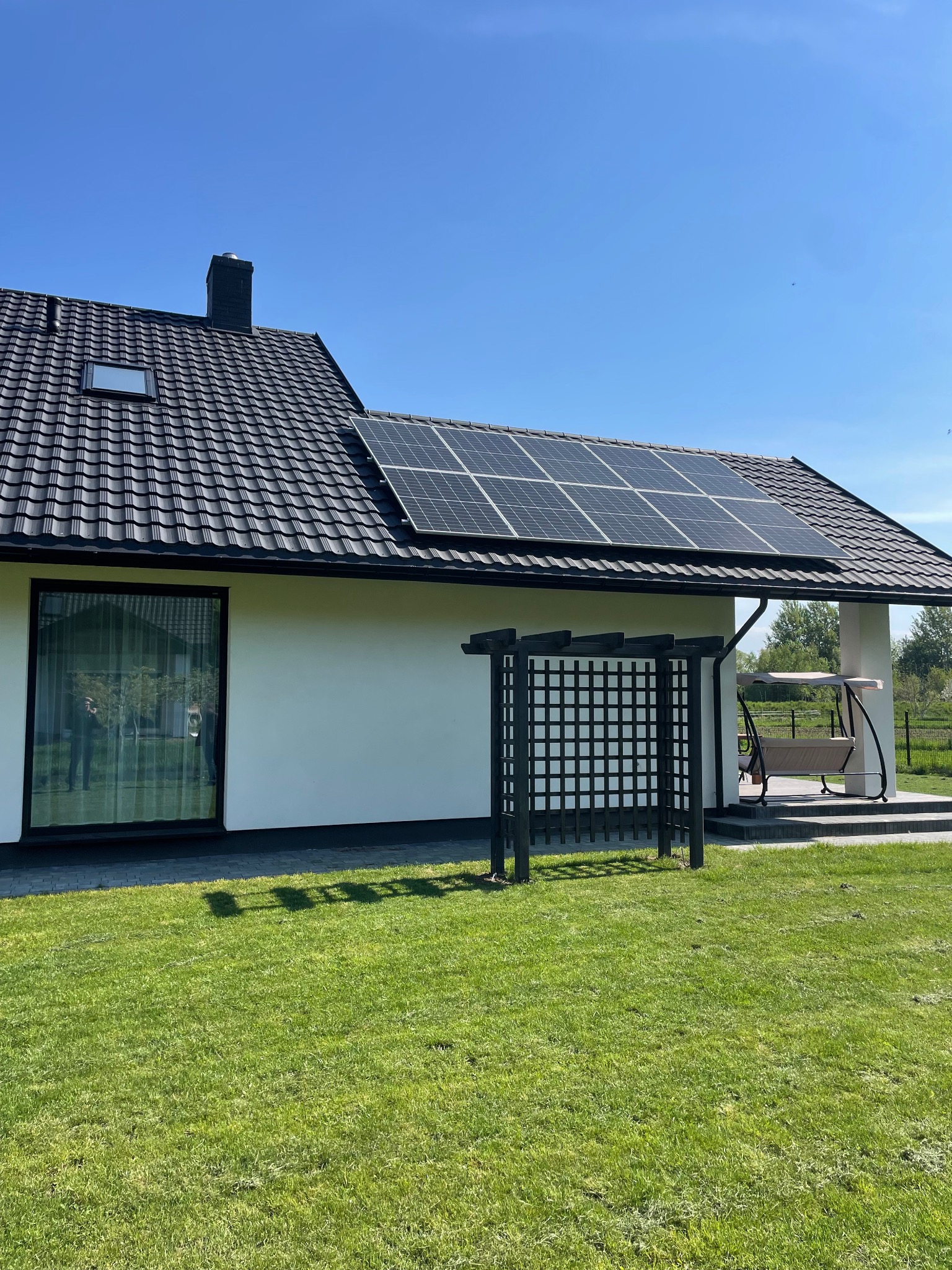 Biały dom jednorodzinny z ciemną dachówką i panelami słonecznymi, widok z ogrodu w słoneczny dzień.