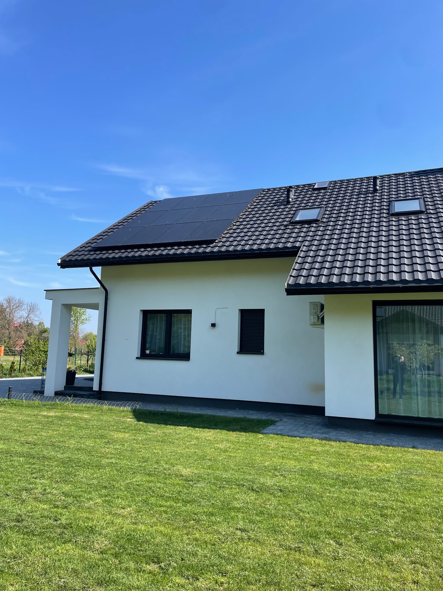 Biały dom jednorodzinny z czarną dachówką i panelami słonecznymi na dachu, widok z poziomu trawnika w słoneczny dzień.