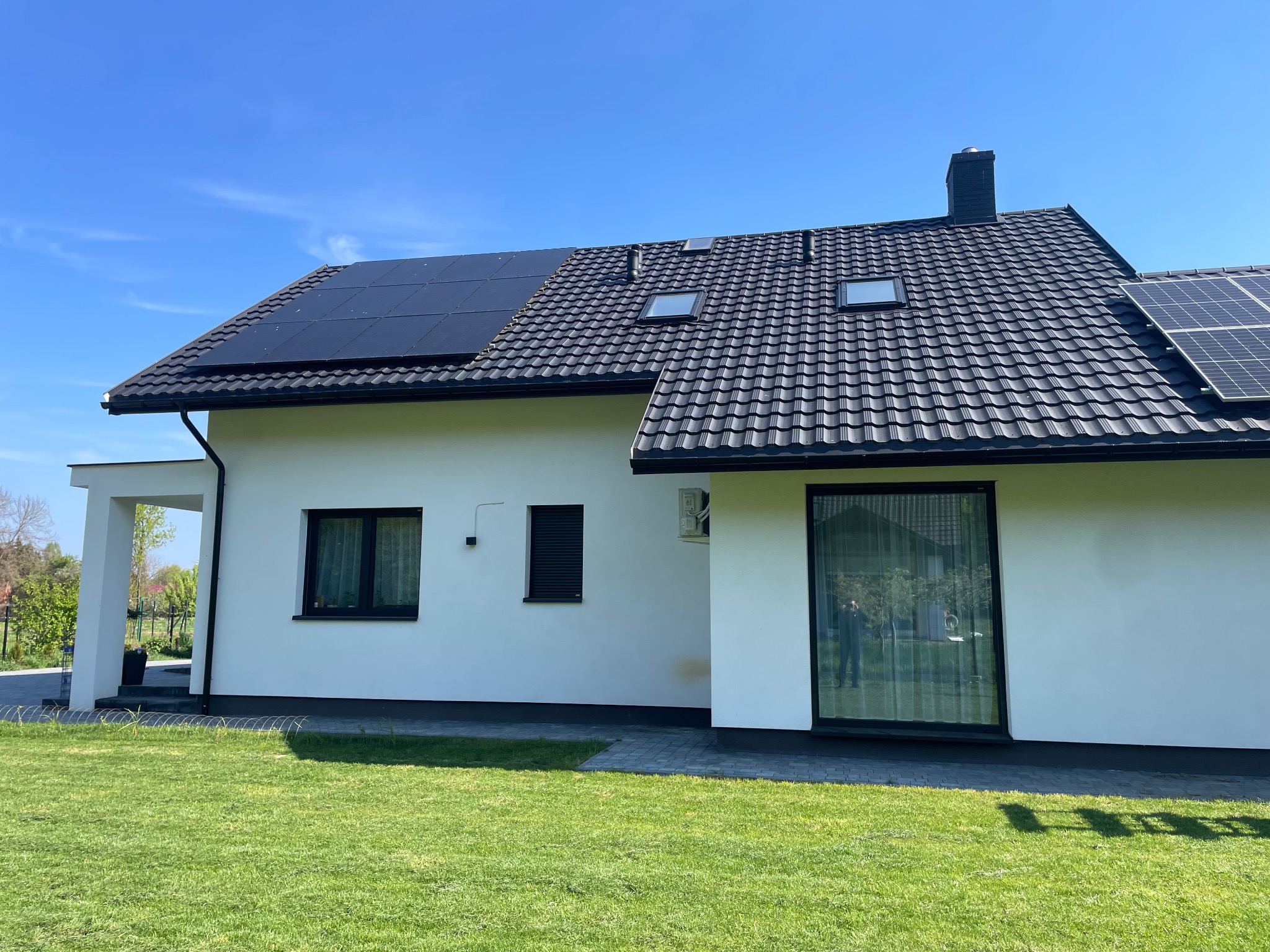 Nowoczesny dom jednorodzinny z białymi ścianami, czarną dachówką, panelami słonecznymi na dachu i zadbanym trawnikiem w słoneczny dzień.