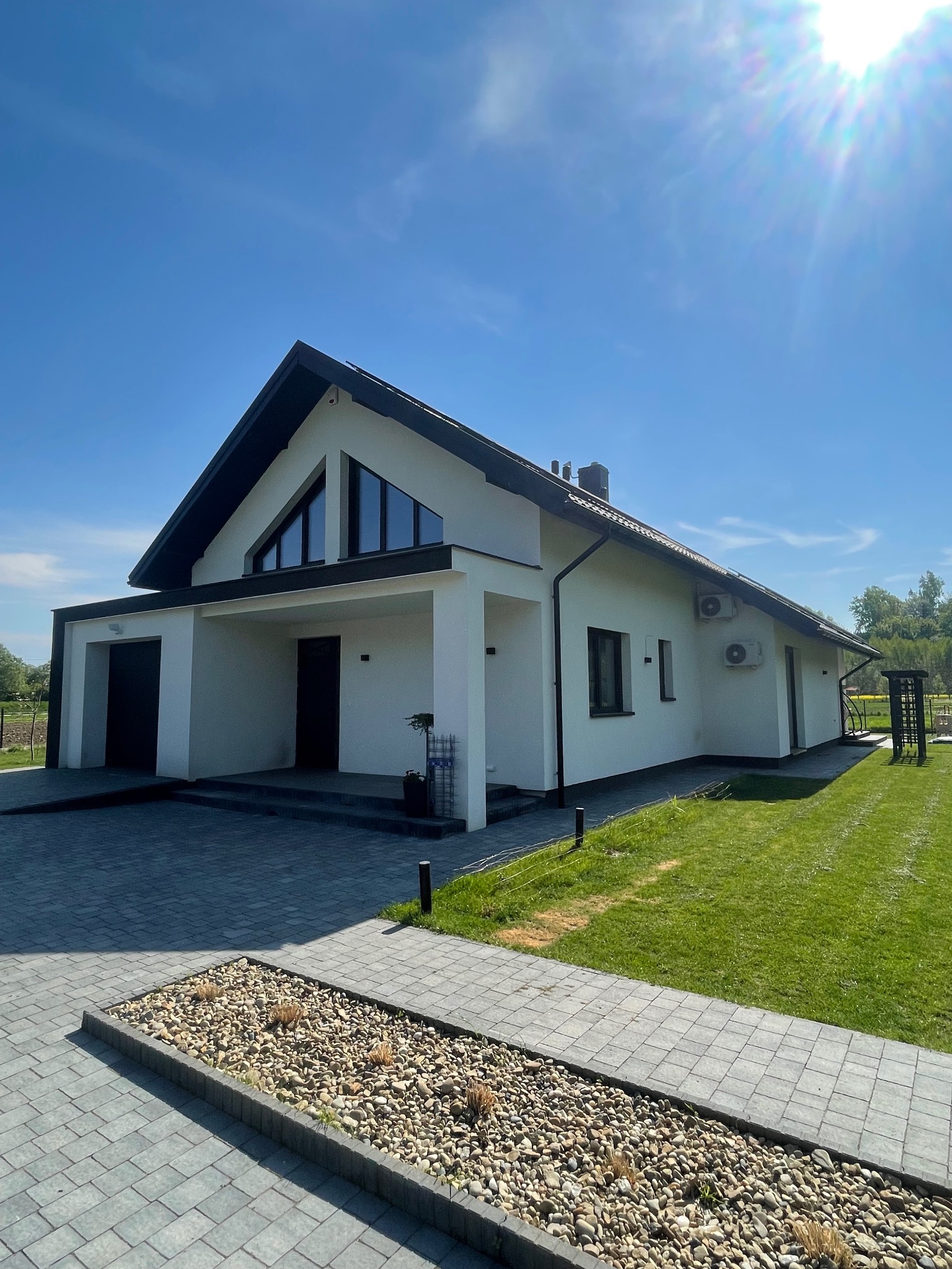 Nowoczesny dom jednorodzinny z białymi ścianami, czarnym dachem i stolarką okienną, widoczny z zewnątrz w słoneczny dzień, z zadbanym trawnikiem i ścieżką z kostki brukowej.