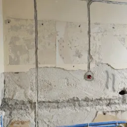 Ściana w trakcie remontu z nową instalacją elektryczną: widoczne kable w peszlach oraz puszka pod gniazdko, ślady po starej farbie i tapetach.