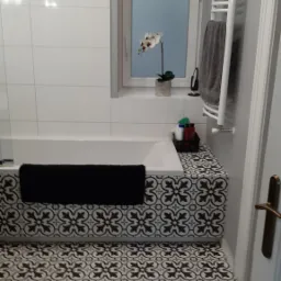 Biała łazienka z wanną obłożoną dekoracyjnymi płytkami, podłoga wyłożona płytkami w marokański wzór, na parapecie okna storczyk w doniczce.