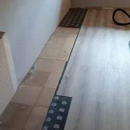 Proces układania paneli podłogowych w pomieszczeniu w Cieszynie, widoczny fragment podkładu i panele o jasnym odcieniu drewna, częściowo ułożone na starej, brudnej podłodze z płytek.