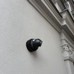 Systemy Zabezpieczen Satklinika - Czarna kamera monitoringu zamontowana na teksturowanej, jasnej ścianie budynku z dekoracyjnymi elementami architektonicznymi w tle. Niski kąt ujęcia.