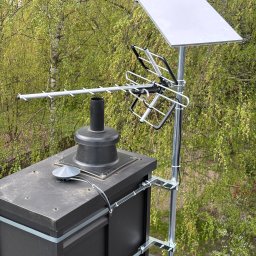 Systemy Zabezpieczen Satklinika - Antena satelitarna i telewizyjna zamontowana na kominie domu, z zielonym tłem drzew. Widoczne kable i elementy mocujące.